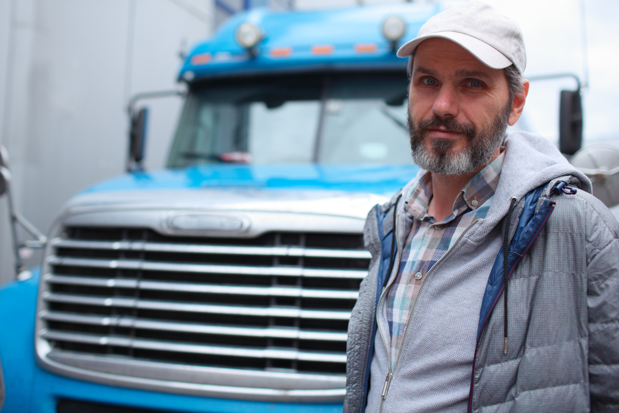A bearded man in warm clothes and a trucker cap stands in front of a large blue truck.
