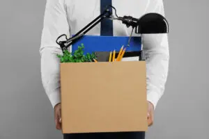 Unemployed man with box of personal office belongings on light grey background, closeup